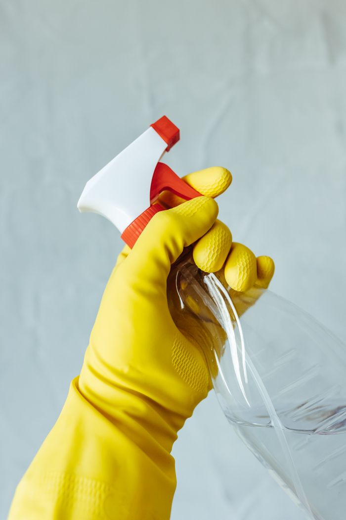 A hand in a yellow rubber glove is holding a red and white spray bottle against a neutral backdrop.
